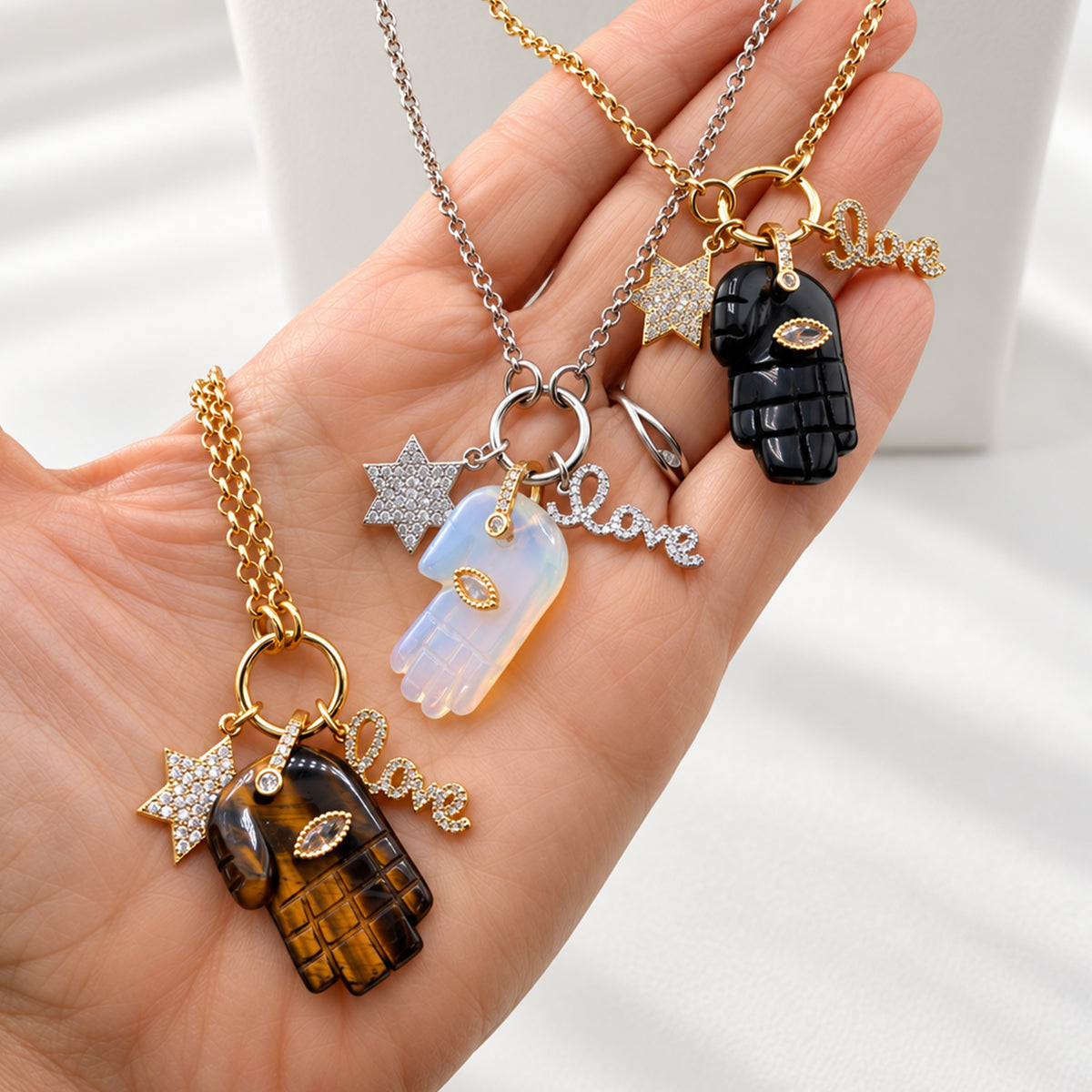 Close-up of Love Lisa Amara necklaces showing gemstone Hamsa pendants, evil eye details, Star of David charms, crystal LOVE charms, and gold-tone chains