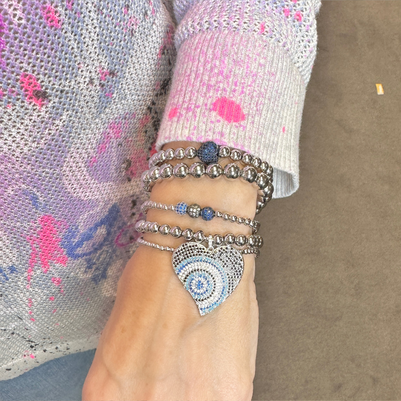 Close-up of a wrist wearing multiple silver bracelets with heart and blue stone designs, against a blurred background.