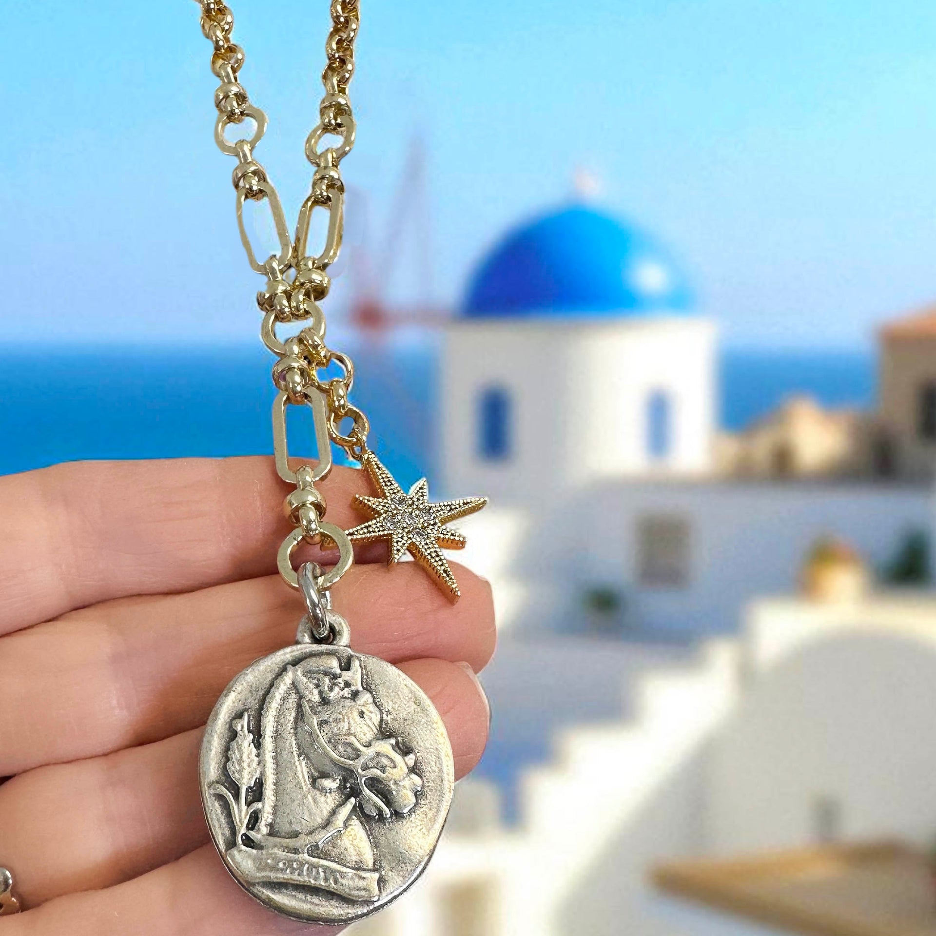 Necklace with a horse pendant held in front of a blue-domed building in Santorini, Greece.