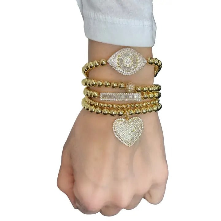Hand wearing multiple gold bracelets with heart and eye designs on a white background