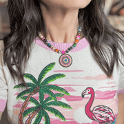 Colorful gemstone fire agate beaded necklace with a round pavé evil eye pendant in pink blue and silver worn on a woman with a palm tree and flamingo print top