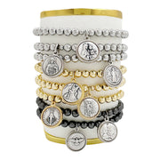 A collection of beaded stretch bracelets with various metallic finish charms representing different saints, displayed on a cylindrical platform.