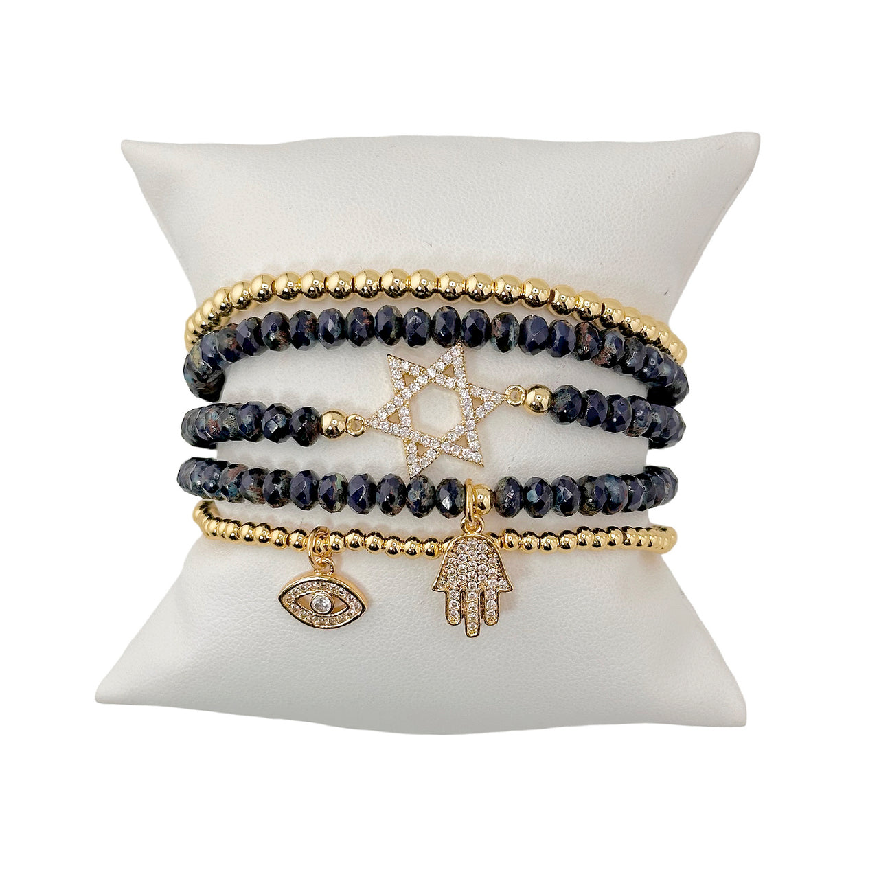 A set of five bracelets displayed on a cushion. The set includes bracelets with charms such as the Star of David, Hand of God, and Evil Eye, featuring gold and dark beads.