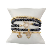 A set of five bracelets displayed on a cushion. The set includes bracelets with charms such as the Star of David, Hand of God, and Evil Eye, featuring gold and dark beads.