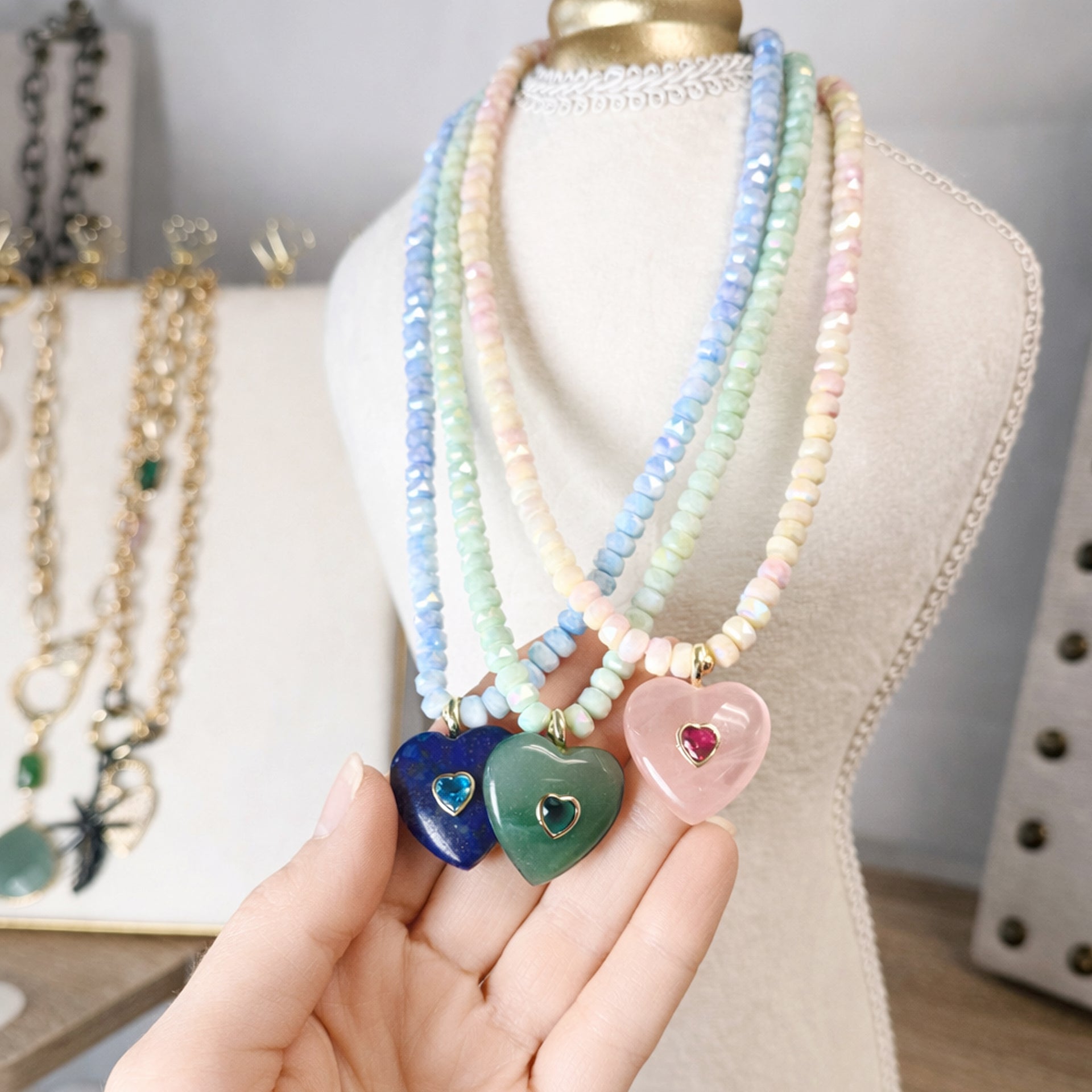 Pastel beaded necklaces with heart-shaped pendants on a mannequin and hand holding one necklace.