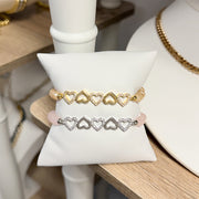 Gold and silver heart-shaped bracelets on a white jewelry display pillow.