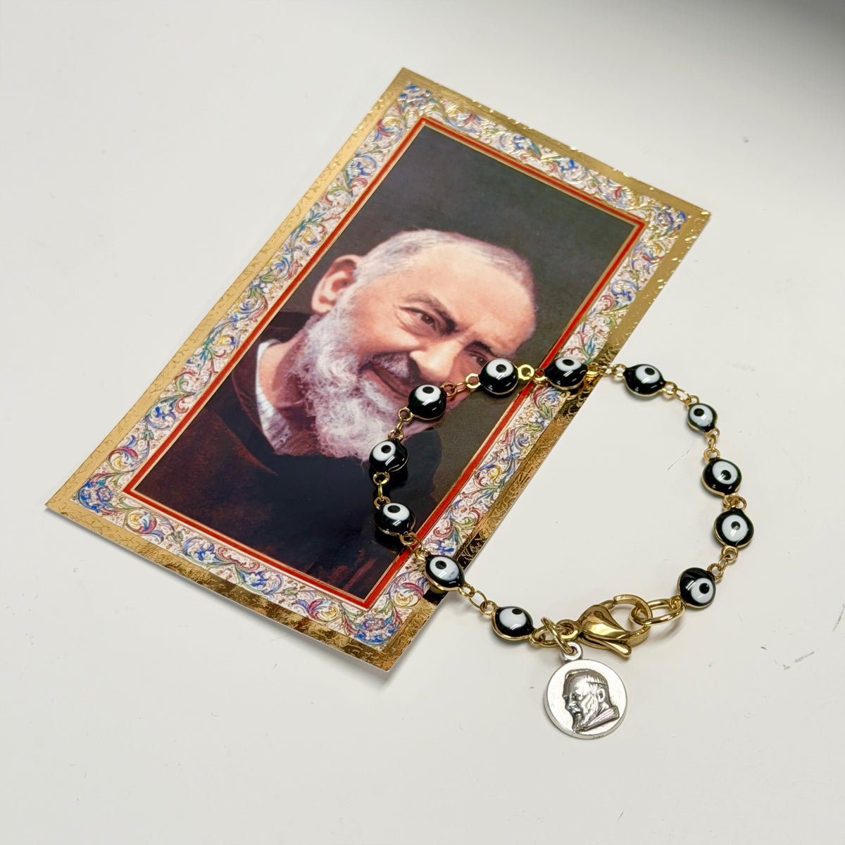 Love Lisa Hannah Evil Eye Saint Medal Bracelet displayed beside its coordinating saint prayer card, showing linked evil eye beads, gold-tone rhodium-plated brass details, and religious medal charm