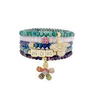 A stack of five colorful bracelets with gold accents and charms, including a glittering bee and a rainbow pavé flower, with each bracelet made of faceted crystal beads.