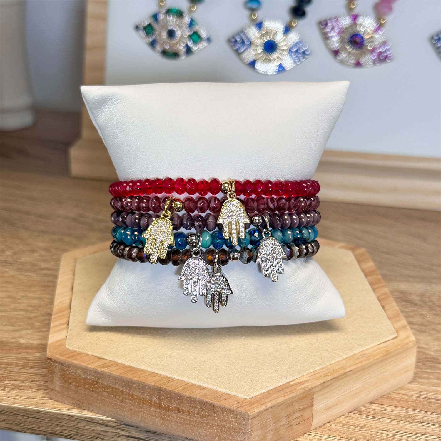 Stack of colorful beaded bracelets with hamsa hand charms on a white pillow, with earrings in the background.