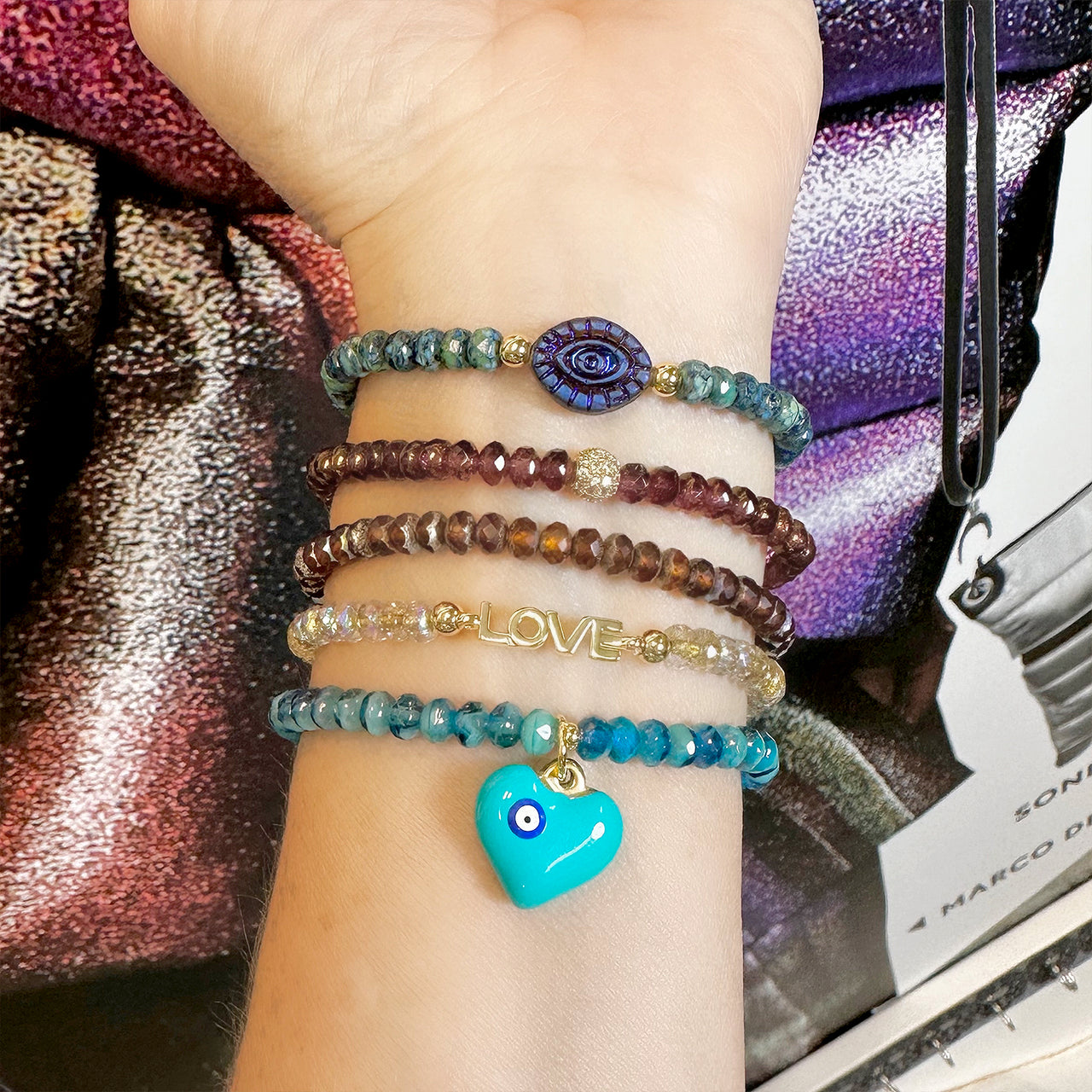 Hand wearing multiple beaded bracelets with a turquoise heart charm on a colorful background