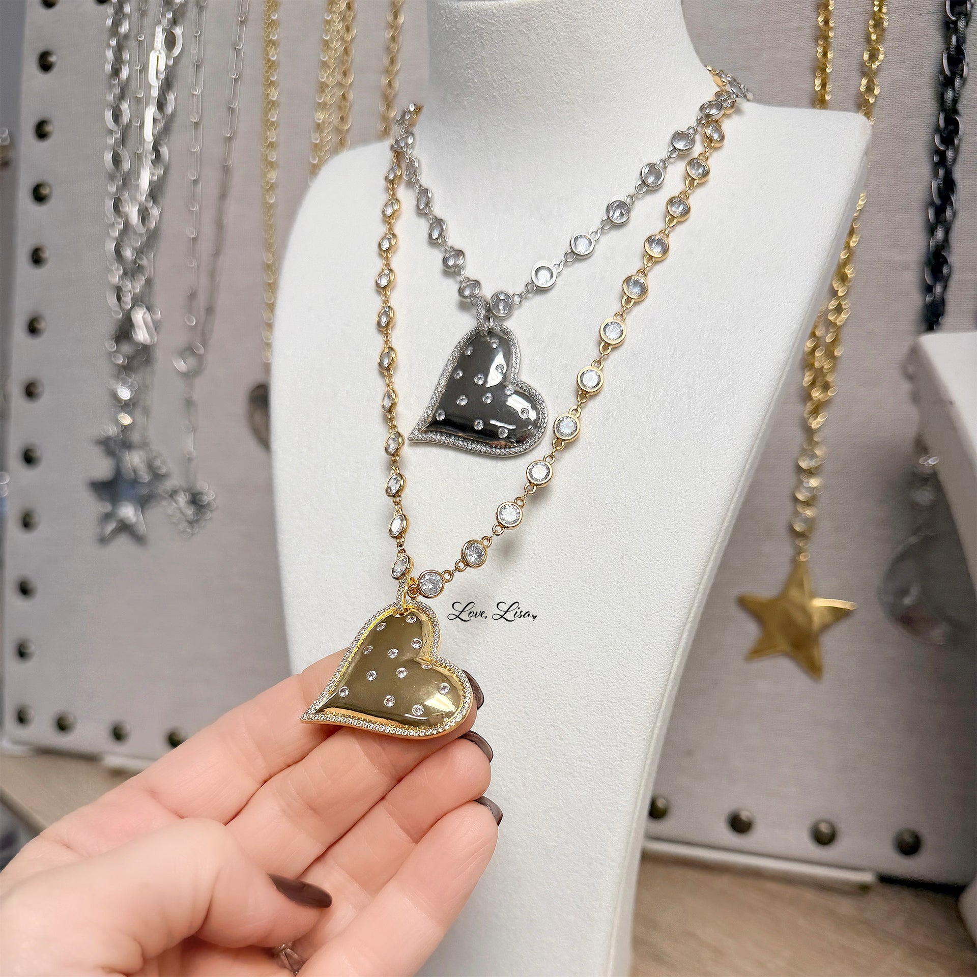 Heart-shaped pendant necklace held by a hand with a display of other necklaces in the background.