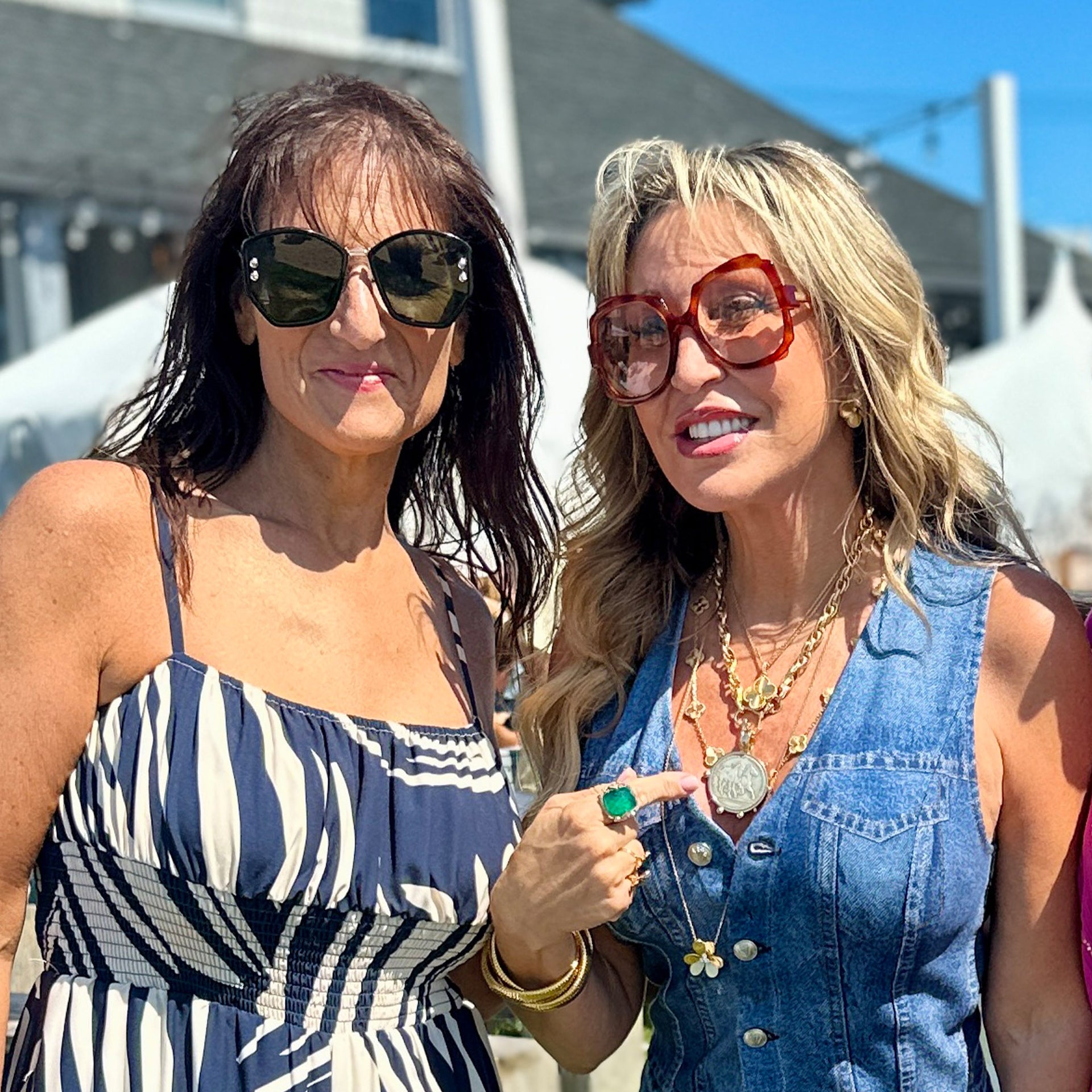 Two women wearing sunglasses outdoors with a clear sky background wearing the horse coin necklace in gold