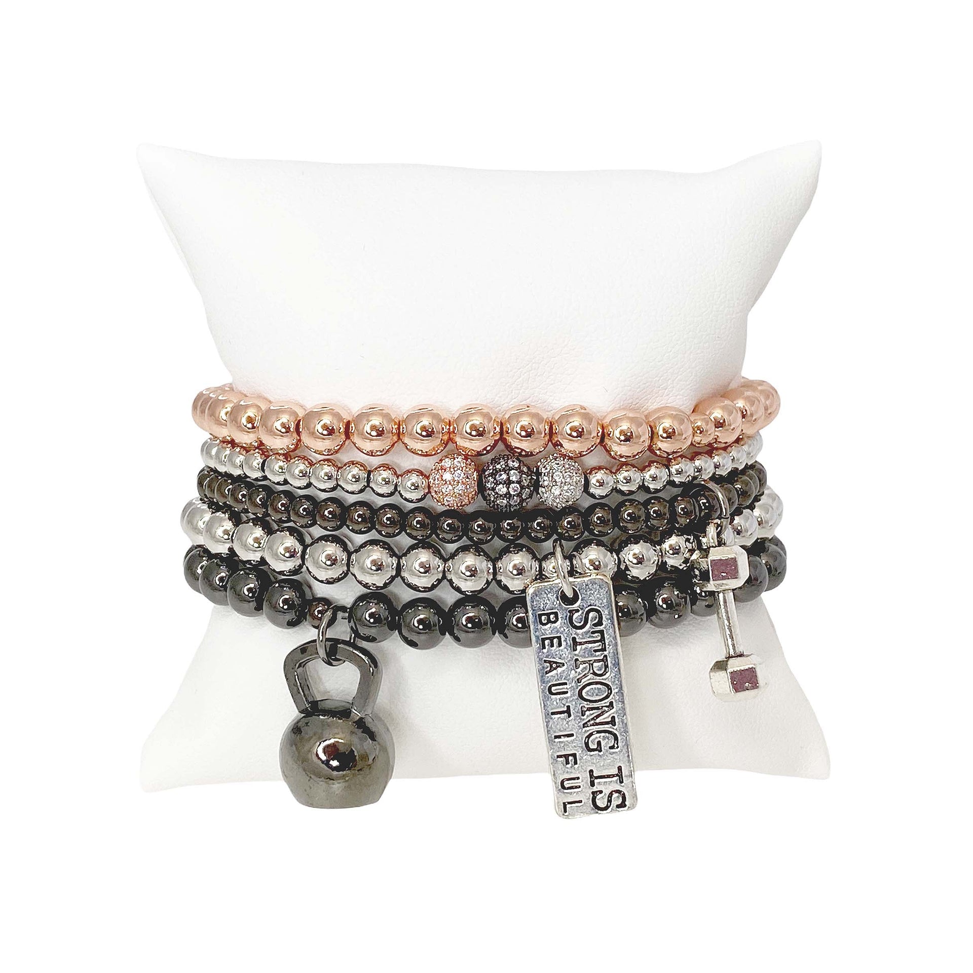 stack of workout inspired bracelets with mixed silver gunmetal and rose gold beads featuring dumbbell kettlebell helmet and Strong Is Beautiful tag on white pillow display
