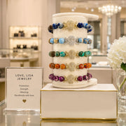 A display stand showcasing five colorful beaded bracelets with silver-toned filigree and gold-toned bead accents, set in a luxury jewelry store next to a sign for 'Love, Lisa Jewelry'.