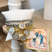 Mary's Blessings 5-piece religious bracelet stack displayed on a white jewelry pillow beside a vintage Marian prayer card. Mixed silver and gold beaded stretch bracelets feature Saint Christopher, Miraculous Mary, Saint Benedict, Holy Spirit Dove, and Guardian Angel charms.