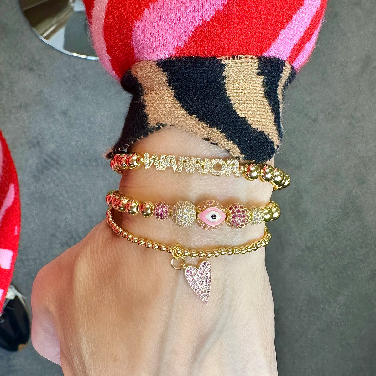 Love Lisa Meghan Breast Cancer Awareness WARRIOR Bracelet Stack worn on wrist with gold-tone beads, pink evil eye detail, pavé heart bracelet, and layered bracelet styling