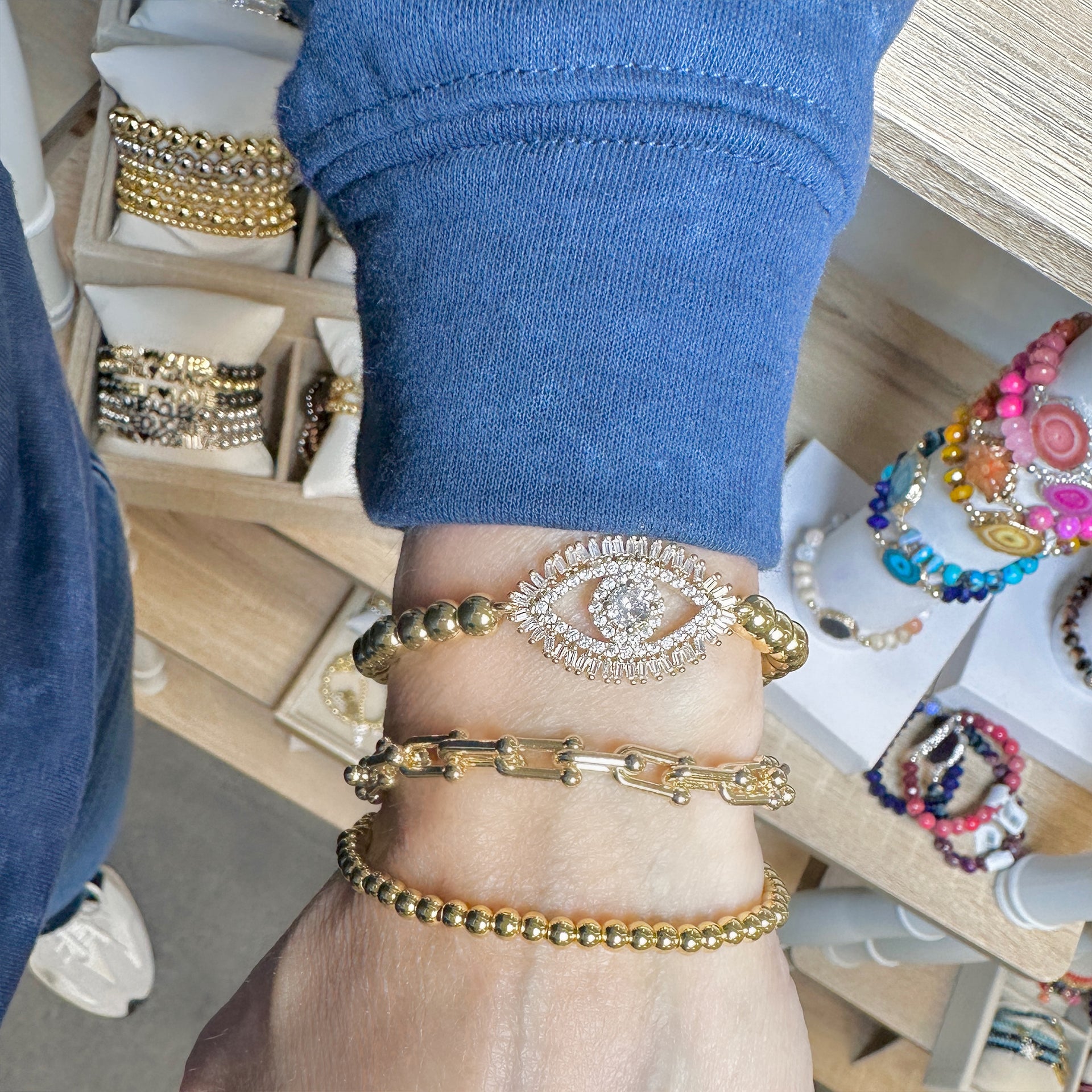 Stacked gold bracelets on wrist, featuring a crystal pavé evil eye centerpiece, the Mila Link gold magnetic clasp bracelet with textured links, and a gold beaded stretch bracelet. Mila Link bracelet also available in silver