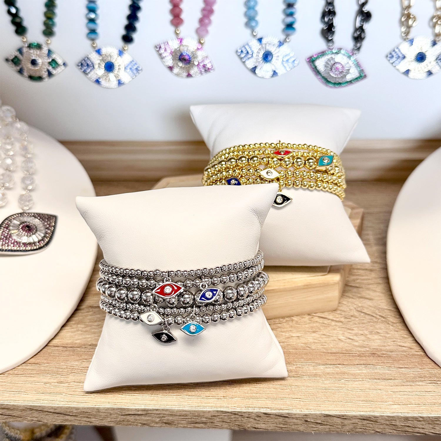 Stack of silver and gold beaded bracelets with colorful enamel evil eye charms displayed on white jewelry pillows in a boutique setting