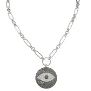 A silver-toned chain necklace featuring a circular evil eye pendant. The pendant is detailed with a textured black outer border, a white center, and a small black pupil, suspended from a polished silver ring on a white background.