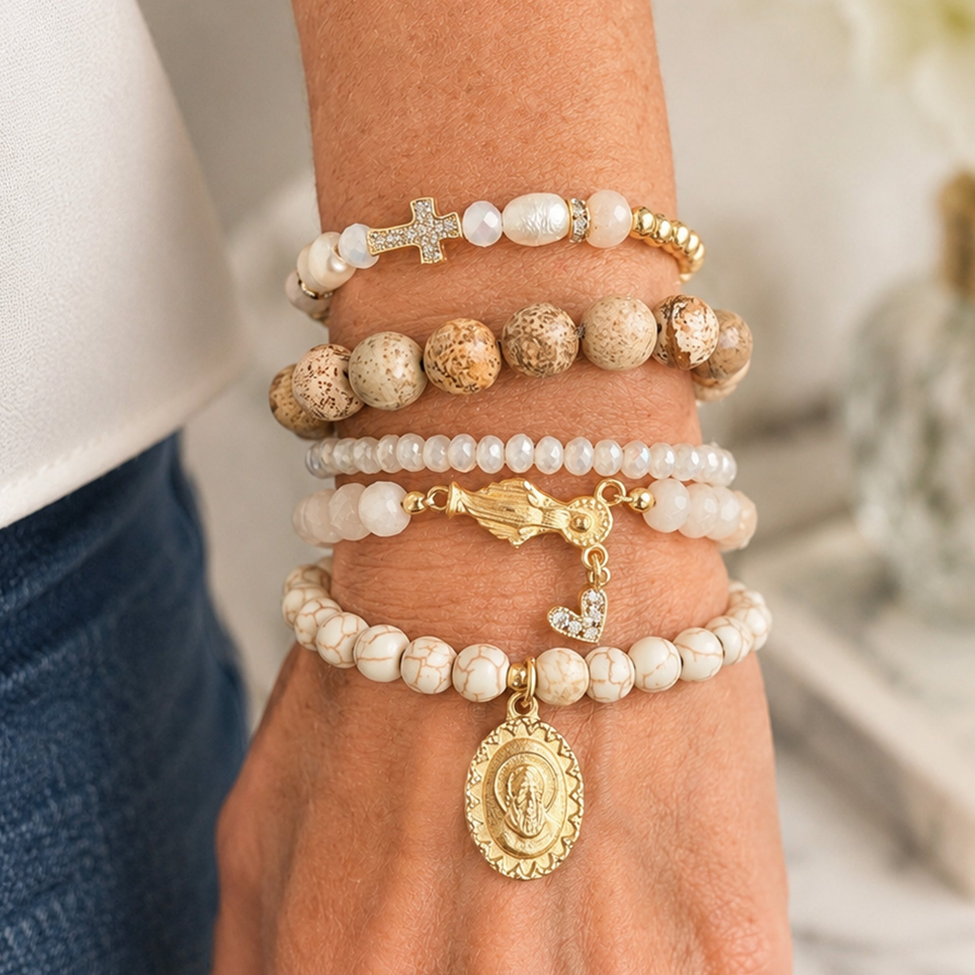A close-up of a person's wrist wearing five stacked beaded bracelets. The bracelets feature white and beige marbled beads, white pearl-like beads, textured brown stone beads, a gold cross charm, a gold bird charm, and a gold oval religious medallion.Rita and Ariana Padre Pio Blessing Bracelet Stack worn on wrist with neutral gemstone bracelets pearls gold accents and religious charms