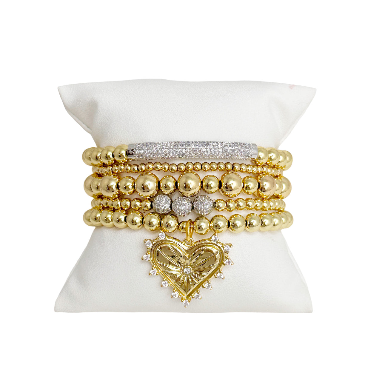 A stack of gold-colored bracelets featuring heart charms and crystal embellishments, displayed on a white pillow.