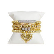 A stack of gold-colored bracelets featuring heart charms and crystal embellishments, displayed on a white pillow.