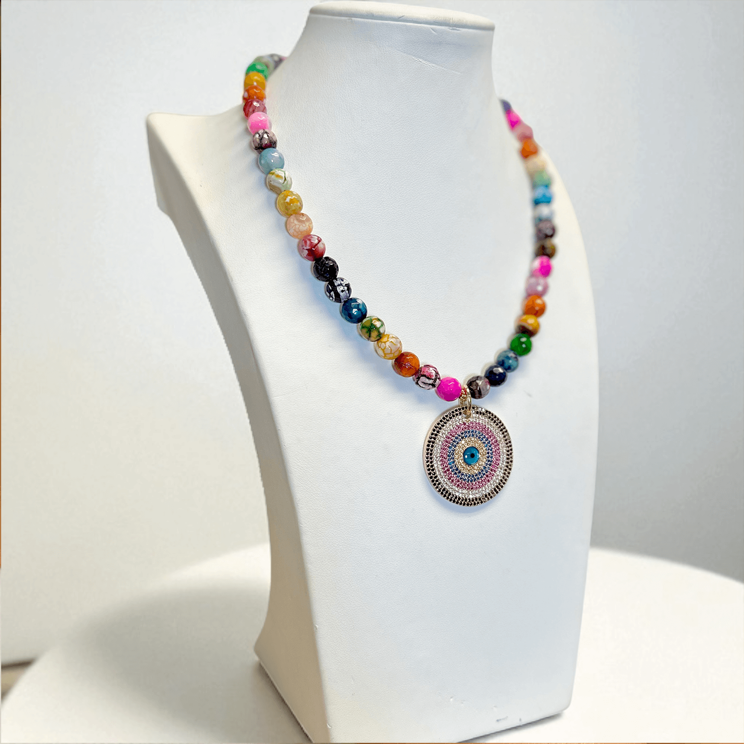Colorful fire agate gemstone beaded necklace with a round pavé evil eye pendant in pink blue and silver displayed on a white jewelry bust