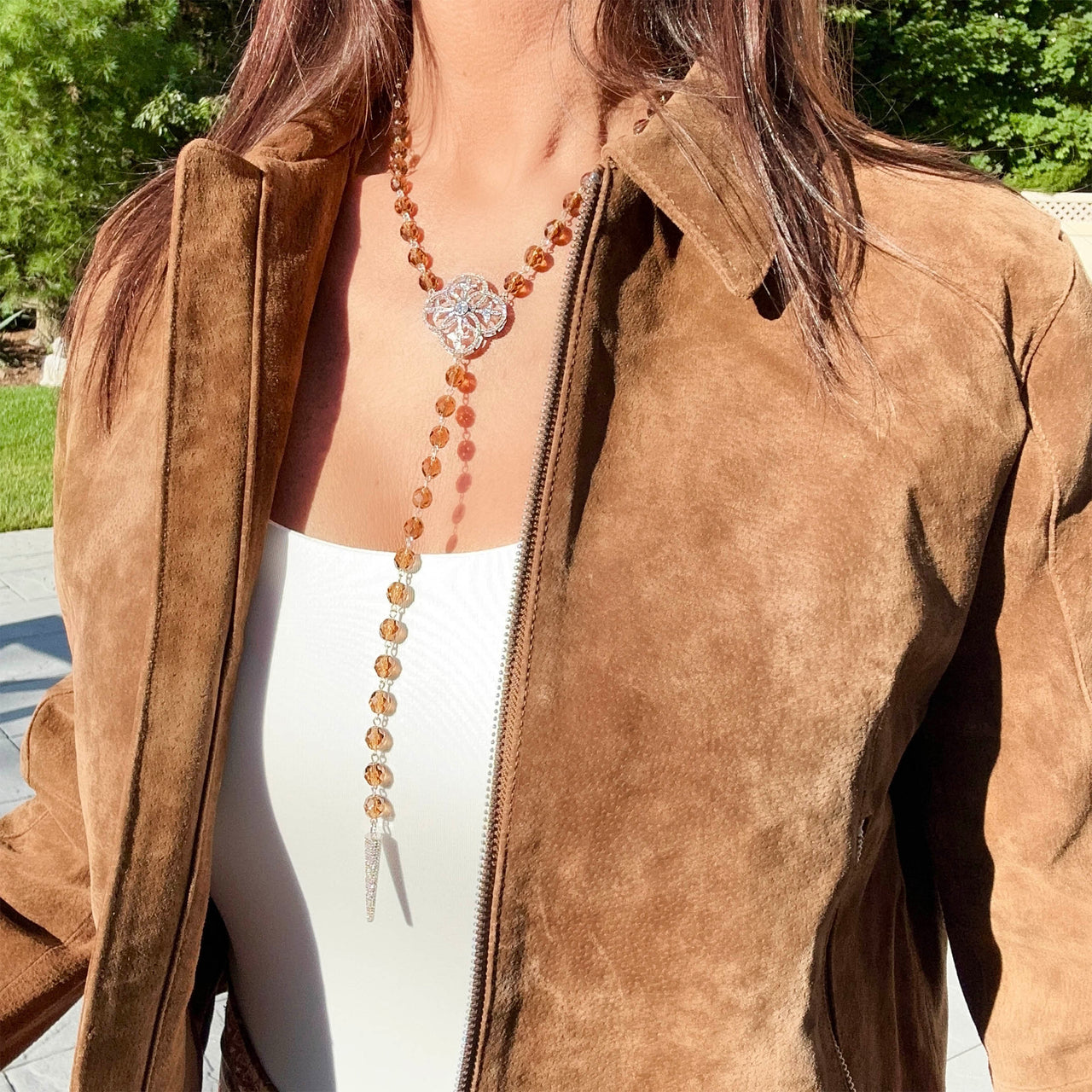 Person wearing a brown suede jacket with a long necklace featuring a decorative pendant.