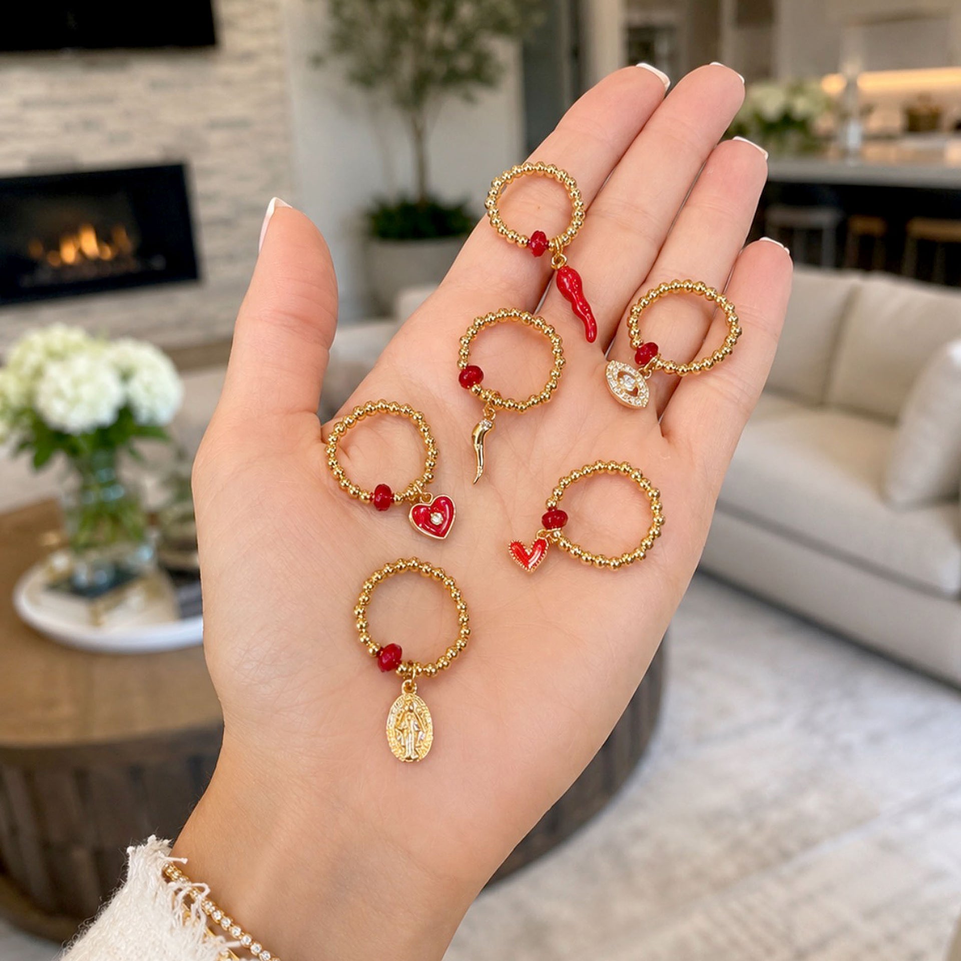 A hand holds several small gold-toned ring-shaped earrings with red heart and bead details, shown against a softly blurred living room background.