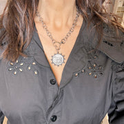 Person wearing a gray blouse with floral embroidery and a necklace with a circular pendant.