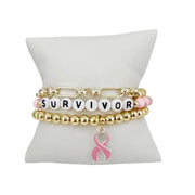 Love Lisa Survivor Breast Cancer Awareness Bracelet Stack in gold displayed on white pillow with SURVIVOR bead detail and dangling pink enamel ribbon charm