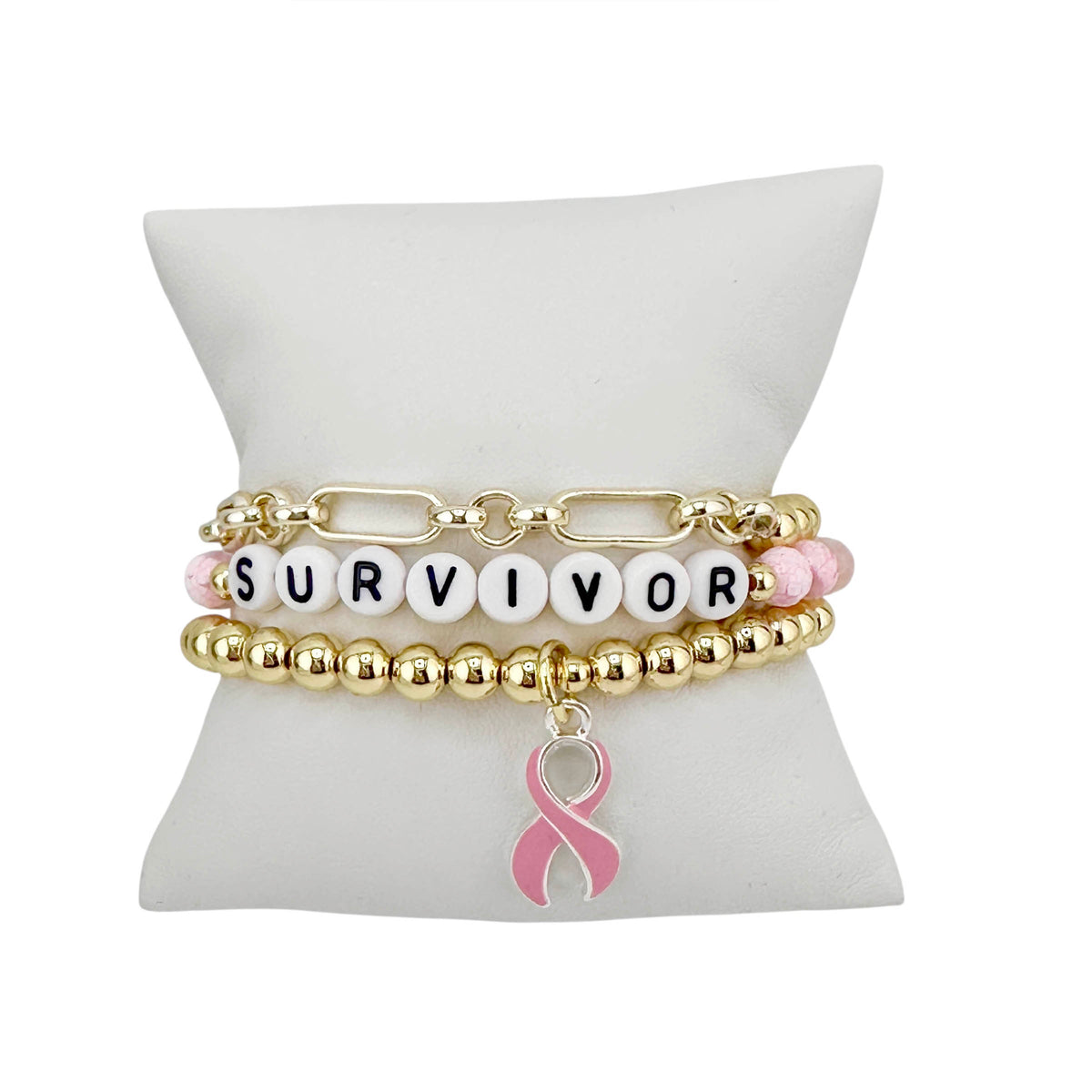 Love Lisa Survivor Breast Cancer Awareness Bracelet Stack in gold displayed on white pillow with SURVIVOR bead detail and dangling pink enamel ribbon charm