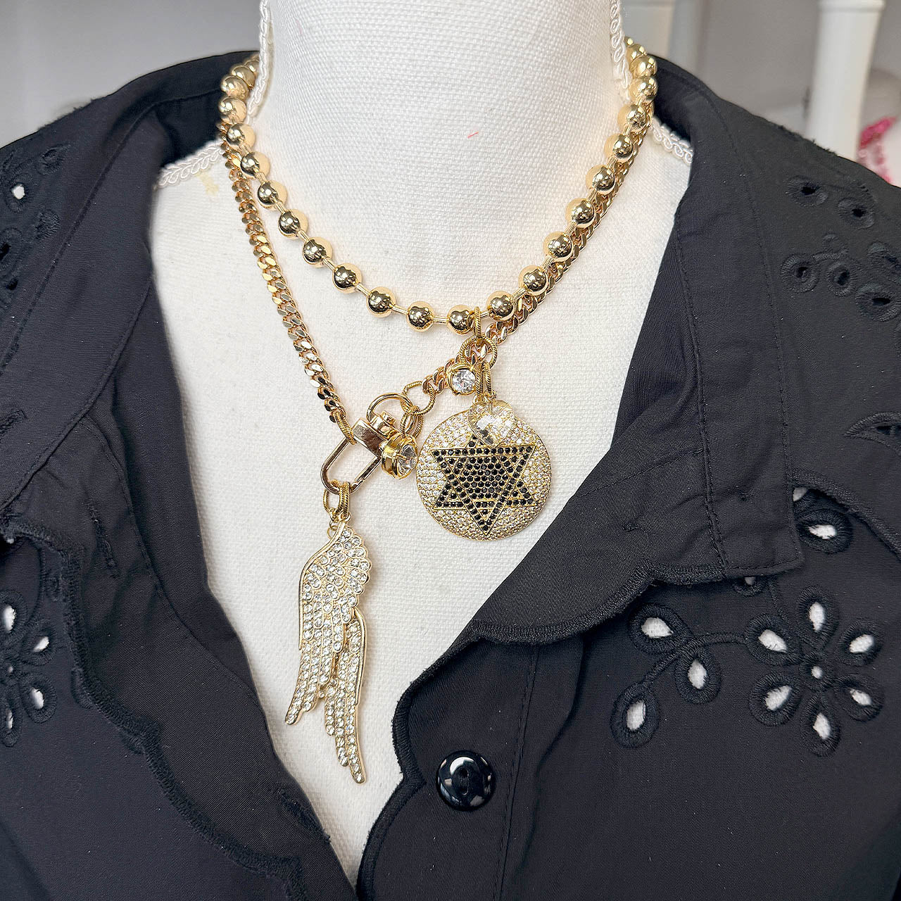 Gold necklace with star of david medallion layered with a necklace with angel wing charms on a mannequin bust wearing a black shirt.