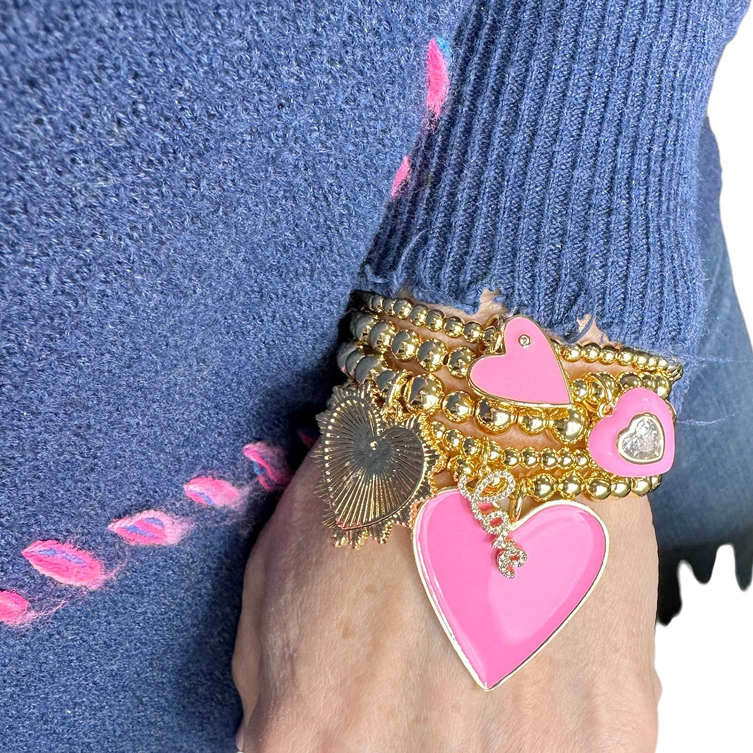 Pink enamel heart bracelet stack worn on wrist featuring gold beaded stretch bracelets