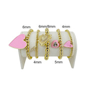 Set of gold bracelets with pink heart charms on a white cylindrical stand, showing size measurements.