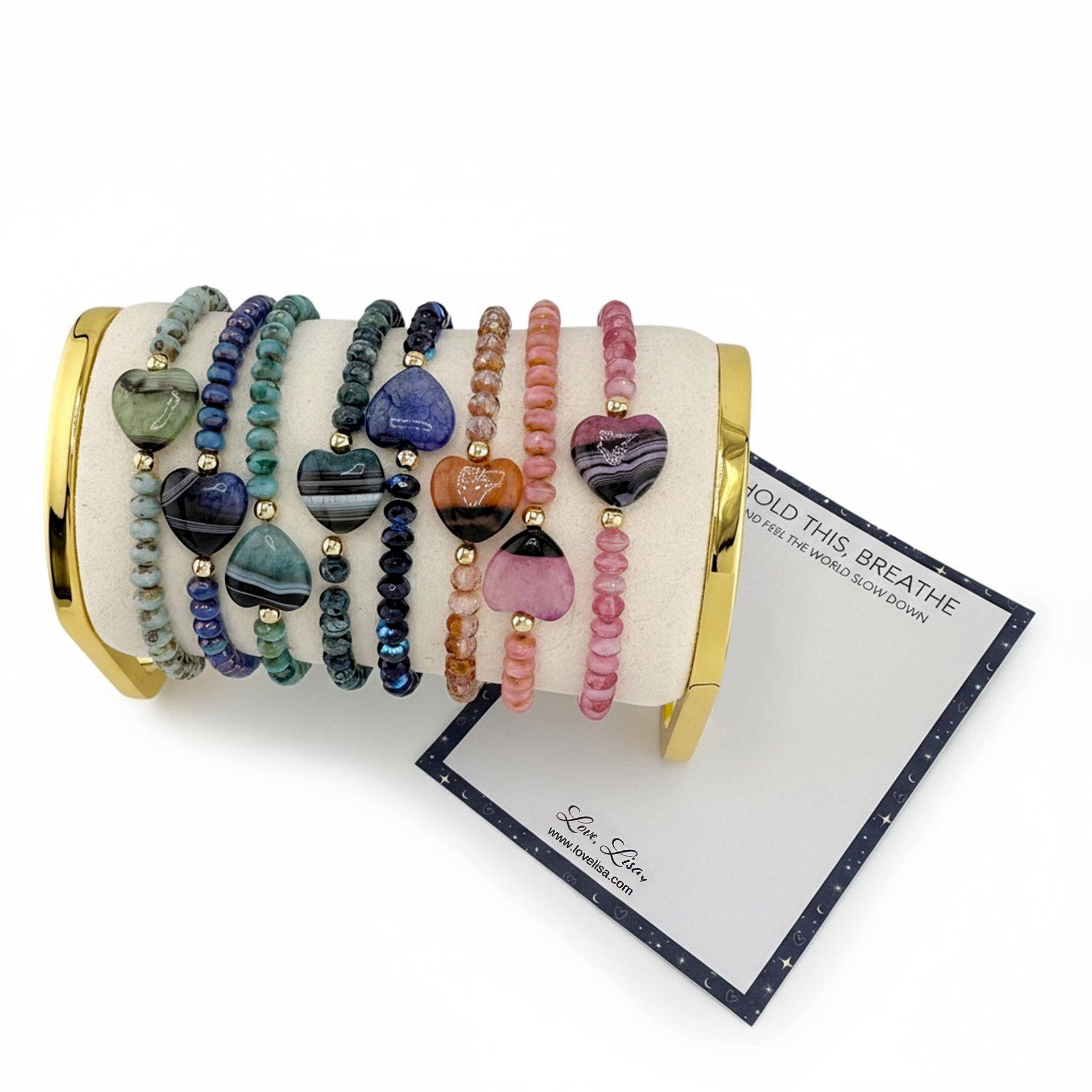 Set of colorful beaded bracelets with heart worry stone on a white cylindrical holder with a gold rim, on a white background.