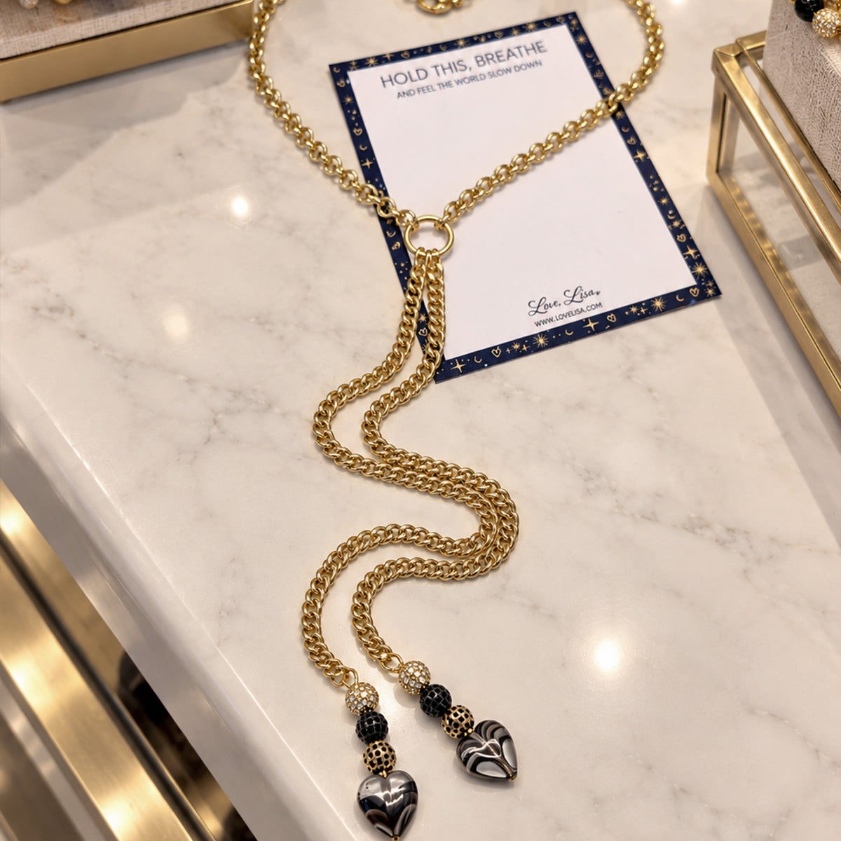 A gold chain necklace with a long wavy section, a small ring connector, and two heart charms, displayed on a white marble surface with a branded card underneath.